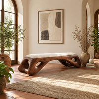 Cushioned bench with a sculpted wood arch base and oval cutouts in a bright entryway with arched windows and plants