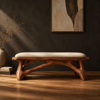 Padded bench with a pale seat on sculpted branching wood legs in a dark studio against a charcoal plaster wall