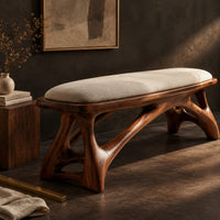 Corner detail of padded bench with a pale seat on sculpted branching wood legs beside wood, brass and linen samples