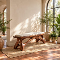 Padded bench with a pale seat on sculpted branching wood legs in a bright entryway with arched windows and plants