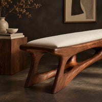 Corner detail of cushioned bench with a carved open frame and slanted legs beside wood, brass and linen samples