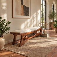 Cushioned bench with a carved open frame and slanted legs in a bright entryway with arched windows and plants