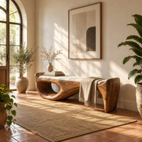 Cushioned bench with a sweeping sculpted body and an oval cutout in a bright entryway with arched windows and plants