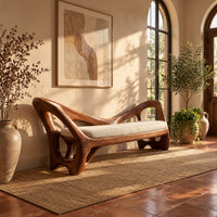 Bench with a cream seat cushion and a sculpted frame full of open cutouts in a bright entryway with arched windows and plants