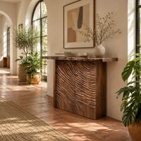 Wooden console table with a live edge top and a deeply carved front in a bright entryway with arched windows and plants