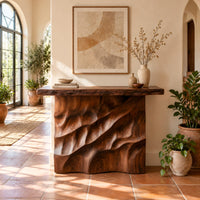Console table with deep diagonal ripple carving on a solid base in a bright entryway with arched windows and plants