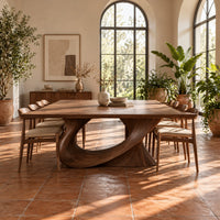 Dining table on a single sweeping pedestal with an oval opening in a bright dining room with arched windows and plants