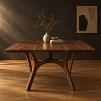 Rectangular dining table with splayed branching legs and an arched cross brace in a dark studio against a charcoal plaster wall