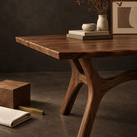 Corner detail of rectangular dining table with splayed branching legs and an arched cross brace beside wood, brass and linen samples