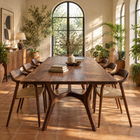 Rectangular dining table with splayed branching legs and an arched cross brace in a bright dining room with arched windows and plants