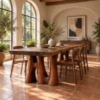 Dining table with a thick slab top on trunk shaped legs with arched openings in a bright dining room with arched windows and plants