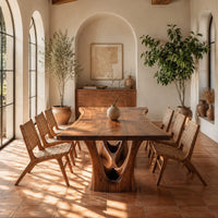 Dining table with a flared wavy top and a carved arch pedestal in a bright dining room with arched windows and plants