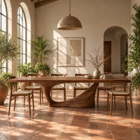 Long dining table on a single swept pedestal with an open arch in a bright dining room with arched windows and plants