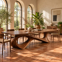 Long dining table on a sweeping ribbon base with one large oval opening in a bright dining room with arched windows and plants