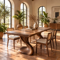 Dining table with two sculpted pedestal legs and a thick flat top in a bright dining room with arched windows and plants