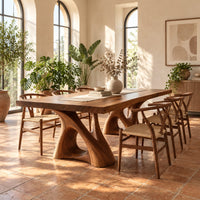 Dining table with a thick plank top on two sculpted loop pedestals in a bright dining room with arched windows and plants