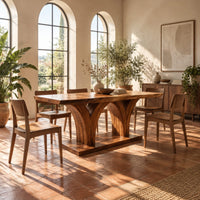 Rectangular dining table with two flared pedestals on a shared low plinth in a bright dining room with arched windows and plants