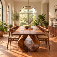 Rectangular dining table on two sculpted legs that form a teardrop gap in a bright dining room with arched windows and plants