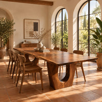Long dining table on two heavy pedestals with arched openings in a bright dining room with arched windows and plants