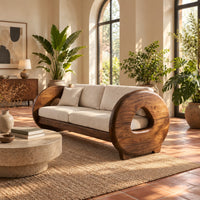 Sofa with thick circular wooden arms, hollow cutouts and cream seat cushions in a bright living room with arched windows and plants