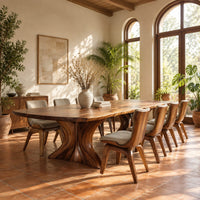 Dining table with a long top and two flared sculpted pedestals in a bright dining room with arched windows and plants