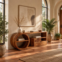 Console table with a drum shaped shelf end, two drawers and an open cubby in a bright entryway with arched windows and plants