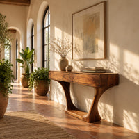 Console table with two flat front panels, flared legs and an oval base in a bright entryway with arched windows and plants