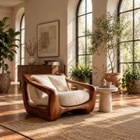 Wooden lounge chair with open sculpted arms and a cushioned seat in a bright living room with arched windows and plants