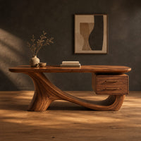 Wooden desk with a flowing sculpted base and two drawers under one end in a dark studio against a charcoal plaster wall