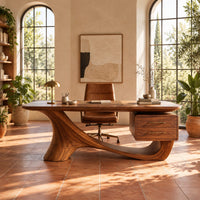 Wooden desk with a flowing sculpted base and two drawers under one end in a bright home office with arched windows and plants