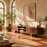 Low media stand with fluted ends, grain front panels and thick cylinder feet in a bright living room with arched windows and plants