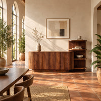 Long fluted sideboard with curved ends and an open shelf tower at one end in a bright dining room with arched windows and plants