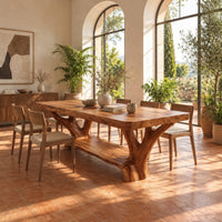 Rectangular dining table on two forked trunk legs joined by a low beam in a bright dining room with arched windows and plants