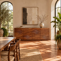 Four door sideboard with carved flowing wave lines on a plinth base in a bright dining room with arched windows and plants