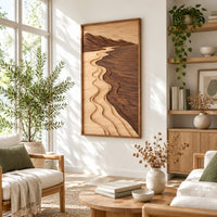 Framed wall art showing a carved river landscape in light and dark wood in a bright sunroom with green plants
