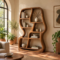 Freeform wooden bookshelf with uneven open cubbies and a soft outline in a bright living room with arched windows and plants