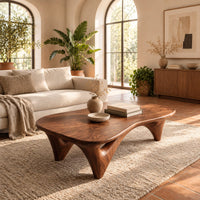 Freeform coffee table with a smooth organic top on sculpted arched legs in a bright living room with arched windows and plants