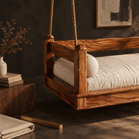 Corner detail of hanging daybed swing with low wood rails and a thick seat pad beside wood, brass and linen samples