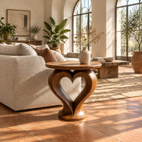Round side table on a sculpted pedestal with a heart shaped opening in a bright living room with arched windows and plants