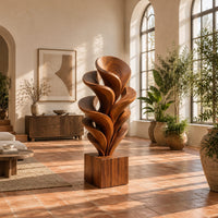 Wood sculpture of layered swirling petals rising from a square block base in a bright living room with arched windows and plants