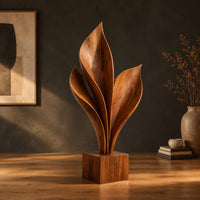 Wood sculpture of sweeping leaf blades rising from a square block base in a dark studio against a charcoal plaster wall