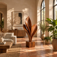Wood sculpture of sweeping leaf blades rising from a square block base in a bright living room with arched windows and plants