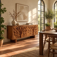 Low sideboard carved with two leafy branch trees across four doors in a bright dining room with arched windows and plants