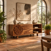 Sideboard with a carved leaf door, open shelf bay and ball feet in a bright dining room with arched windows and plants