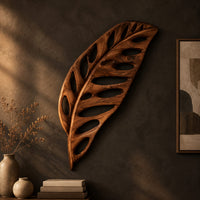 Leaf shaped wooden wall sculpture with open oval cutouts and carved veins on a dark charcoal plaster wall