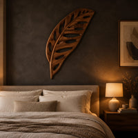 Leaf shaped wooden wall sculpture with open oval cutouts and carved veins above a bed in a dusk lit bedroom
