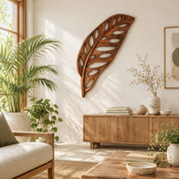 Leaf shaped wooden wall sculpture with open oval cutouts and carved veins in a bright sunroom with green plants