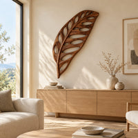 Leaf shaped wooden wall sculpture with open oval cutouts and carved veins above a light oak sideboard in daylight
