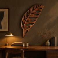 Leaf shaped wooden wall sculpture with open oval cutouts and carved veins above a desk in a dark study corner