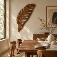 Leaf shaped wooden wall sculpture with open oval cutouts and carved veins beside a dining table in natural light